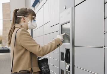 Woman wearing face mask and gloves using parcel terminal - AHSF02448