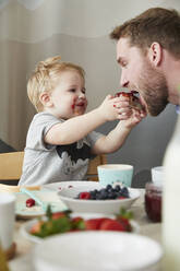 Porträt eines verschmierten kleinen Jungen, der seinen Vater am Frühstückstisch mit Marmeladenbrot füttert - FSF01054