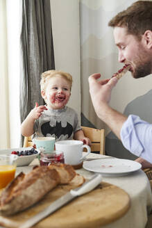 Porträt eines verschmierten kleinen Jungen am Frühstückstisch, der seinen Vater beim Essen von Brot mit Marmelade beobachtet - FSF01058