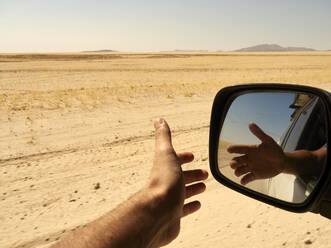 Reflection in the rear view mirror of a man's hand outside the window while driving in the desert, Sossusvlei, Namibia. - VEGF02086