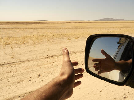 Reflexion der Hand eines Mannes vor dem Fenster im Rückspiegel während einer Fahrt durch die Wüste, Sossusvlei, Namibia. - VEGF02086