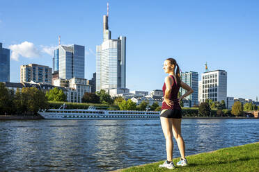 Deutschland, Hessen, Frankfurt, Junge Frau in Sportkleidung am Mainufer stehend mit Stadtsilhouette im Hintergrund - PUF01879