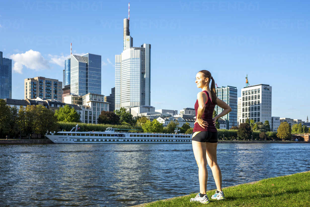 Germany, Hesse, Frankfurt, Young woman in sportswear standing on bank of  Main with city skyline in background stock photo