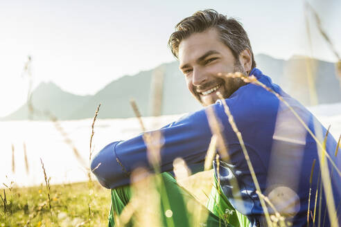 Porträt eines lächelnden Mannes, der in der Morgendämmerung auf einer Bergwiese sitzt, Achenkirch, Österreich - SDAHF00883