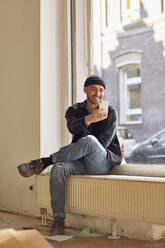Mann renoviert Ladenlokal, sitzt auf der Fensterbank, benutzt Smartphone - MCF00792