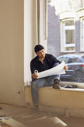 Man refurbishing shop location, sitting on windowsill, studying blueprint - MCF00800