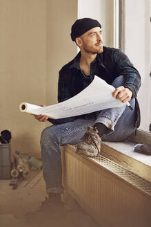 Man refurbishing shop location, sitting on windowsill, studying blueprint - MCF00804