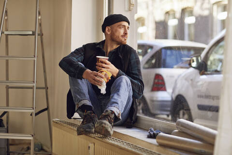 Mann renoviert Ladenlokal, sitzt auf der Fensterbank und trinkt Kaffee, lizenzfreies Stockfoto