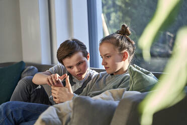Brother and sister sitting on couch at home using smartphone - AUF00359