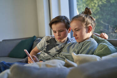 Brother and sister sitting on couch at home using smartphone - AUF00360