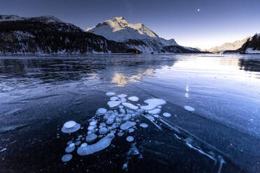 Methanblasen in der eisigen Oberfläche des Sees mit schneebedeckter Spitze, beleuchtet vom Mondlicht, Sils, Engadin, Graubünden, Schweizer Alpen, Schweiz, Europa - RHPLF14840