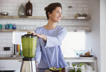 Smiling brunette woman making healthy green smoothie in blender in kitchen - CAIF26968