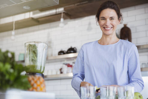 Porträt einer lächelnden, selbstbewussten brünetten Frau, die einen gesunden grünen Smoothie in einem Mixer in der Küche zubereitet - CAIF26970