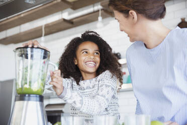 Enthusiastische Tochter hilft ihrer Mutter bei der Zubereitung eines gesunden grünen Smoothies im Mixer in der Küche - CAIF26978