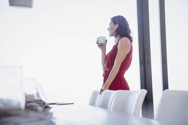 Pensive brunette woman drinking coffee and looking out window in dining room - CAIF26995