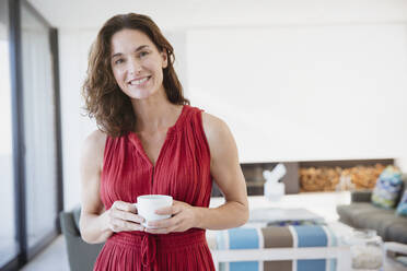 Porträt einer lächelnden, selbstbewussten brünetten Frau, die im Wohnzimmer Kaffee trinkt - CAIF26998
