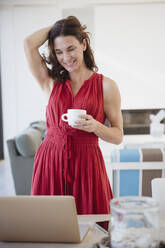 Lächelnde brünette Frau trinkt Kaffee und arbeitet am Laptop im Esszimmer - CAIF27000