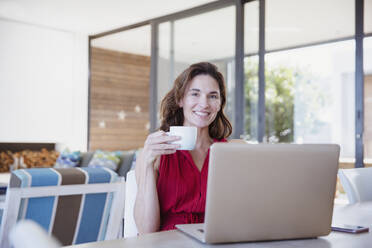 Porträt einer lächelnden, selbstbewussten brünetten Frau, die im Esszimmer Kaffee trinkt und einen Laptop benutzt - CAIF27001