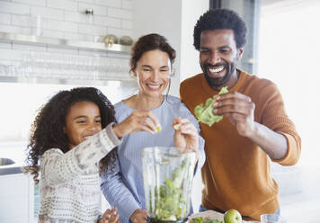 Smiling multi-ethnic family making green smoothie in blender in kitchen - CAIF27009