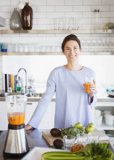 Porträt einer lächelnden, zuversichtlichen brünetten Frau, die gesunden Karottensaft an einem Mixer in der Küche trinkt - CAIF27011