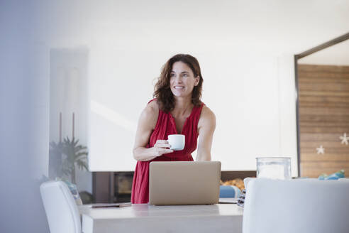 Lächelnde brünette Frau trinkt Kaffee, arbeitet am Laptop am Esstisch - CAIF27022
