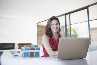 Portrait smiling brunette woman working at laptop in kitchen - CAIF27033