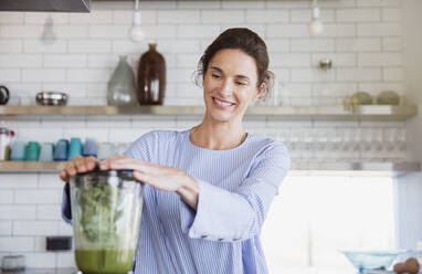 Lächelnde Frau bei der Zubereitung eines gesunden grünen Smoothies in einem Mixer in der Küche - CAIF27045