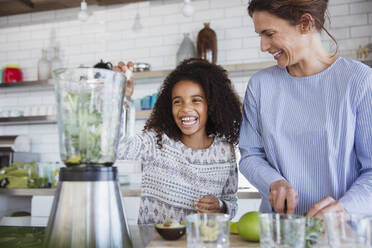 Mother and daughter making healthy green smoothie in blender in kitchen - CAIF27047