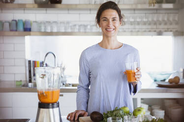 Porträt einer selbstbewussten, lächelnden Frau, die in der Küche gesunden Karottensaft trinkt - CAIF27052