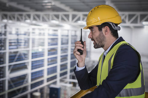 Male supervisor talking, using walkie-talkie on platform in factory - CAIF27059