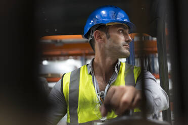 Männlicher Arbeiter fährt Gabelstapler und schaut über die Schulter in einer Fabrik - CAIF27135