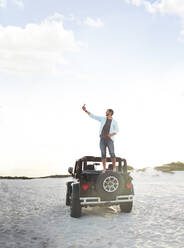 Junger Mann mit Fotohandy, der ein Selfie auf einem Jeep-Reifen am Strand macht - CAIF27167