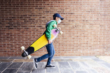 Junge mit Schutzmaske und Longboard vor einer Backsteinmauer - JCMF00682