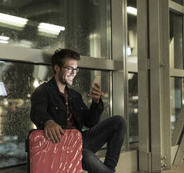 Lächelnder junger Mann mit Koffer sitzt am verregneten Fenster und benutzt sein Smartphone - UUF20304