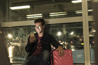 Lächelnder junger Mann mit Koffer sitzt am verregneten Fenster und benutzt sein Smartphone - UUF20305