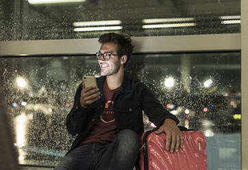 Lächelnder junger Mann mit Koffer sitzt am verregneten Fenster und benutzt sein Smartphone - UUF20306