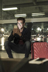 Lächelnder junger Mann mit Koffer sitzt am verregneten Fenster und benutzt sein Smartphone - UUF20307