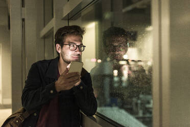 Junger Mann mit Smartphone schaut nachts aus einem verregneten Fenster - UUF20310
