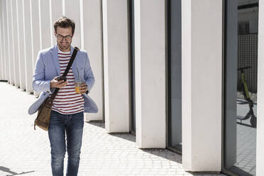 Junger Mann beim Spaziergang in der Stadt mit Handy und Getränk zum Mitnehmen, Lissabon, Portugal - UUF20318