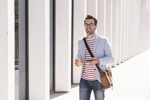 Lächelnder junger Mann in der Stadt mit Handy und Getränk zum Mitnehmen, Lissabon, Portugal - UUF20322