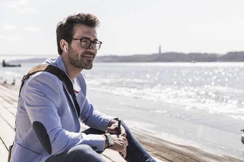 Smiling young man with earbuds sitting on the waterfront stock photo