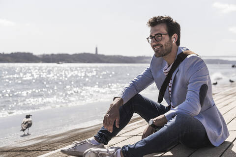 Lächelnder junger Mann mit Ohrstöpseln am Ufer sitzend, lizenzfreies Stockfoto