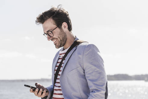 Lächelnder junger Mann mit Smartphone am Wasser - UUF20340