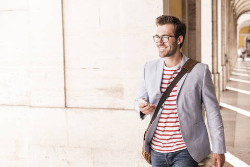 Smiling young man with smartphone in the city, Lisbon, Portugal - UUF20343