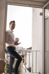 Junger Mann mit Mobiltelefon und Kaffeetasse auf einem Balkon - UUF20354