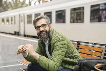 Mann sitzt auf einer Bank am Bahnhof und isst ein Sandwich - AHSF02477