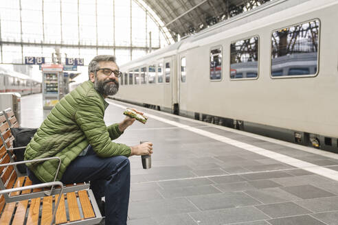 Mann sitzt auf einer Bank am Bahnhof mit Sandwich und heißem Getränk - AHSF02478