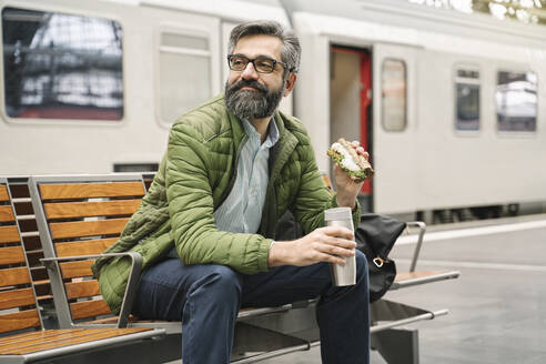 Mann sitzt auf einer Bank am Bahnhof mit Sandwich und heißem Getränk - AHSF02496