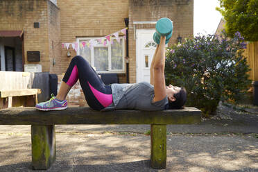 Full length side view of mature woman exercising with dumbbells on wooden bench at back yard - PMF01037