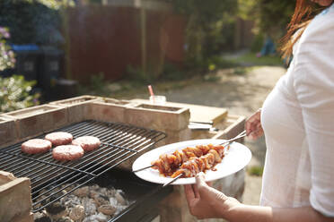 Mittelteil einer Frau, die einen Teller mit Spießen hält, während sie an einem sonnigen Tag am Grill im Hinterhof steht - PMF01045